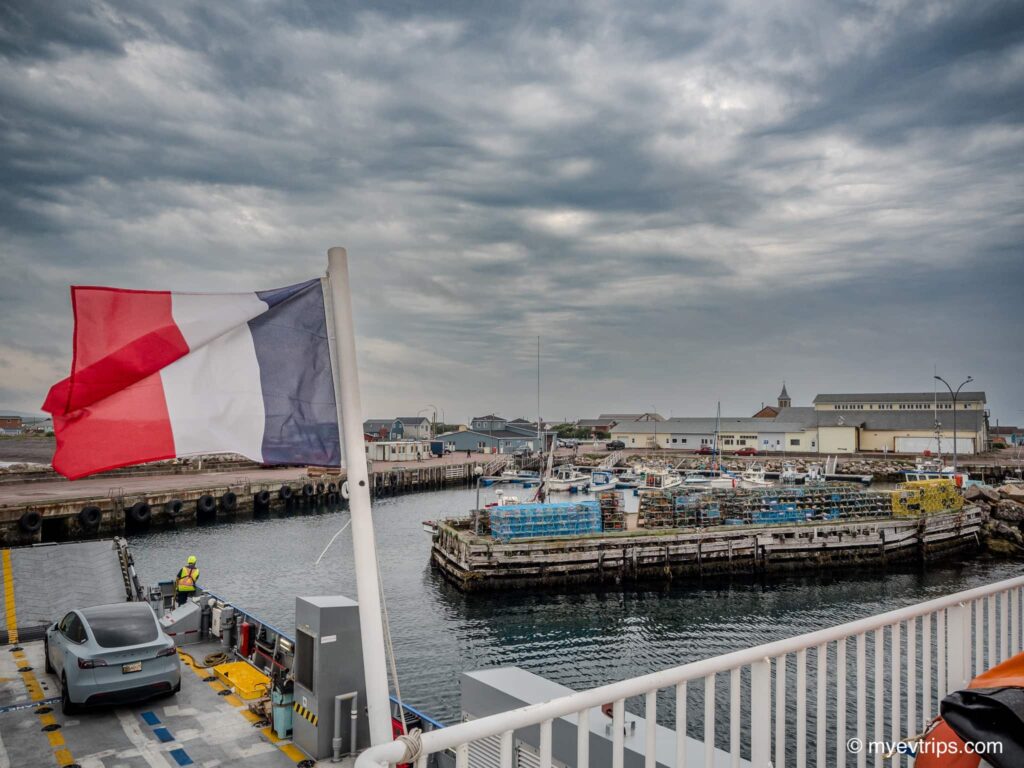 A Tesla Model Y on a Saint Pierre et Miquelon EV road trip ferry to with France flag in foreground 
