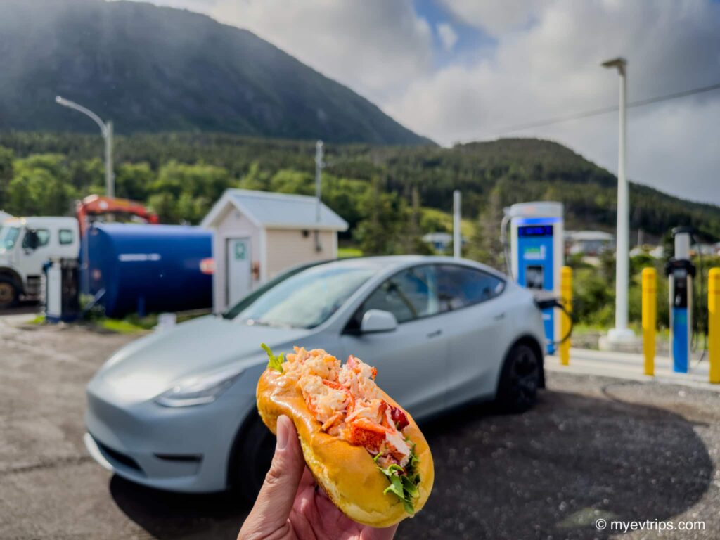 hand holding up lobster roll in front of Tesla Model Y charging on a Newfoundland EV road trip