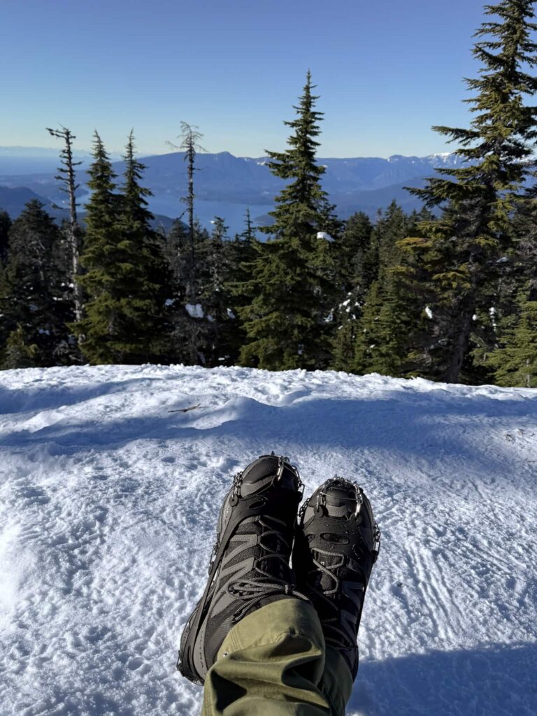 legs with hiking boots on snowy mountain with ocean views in background